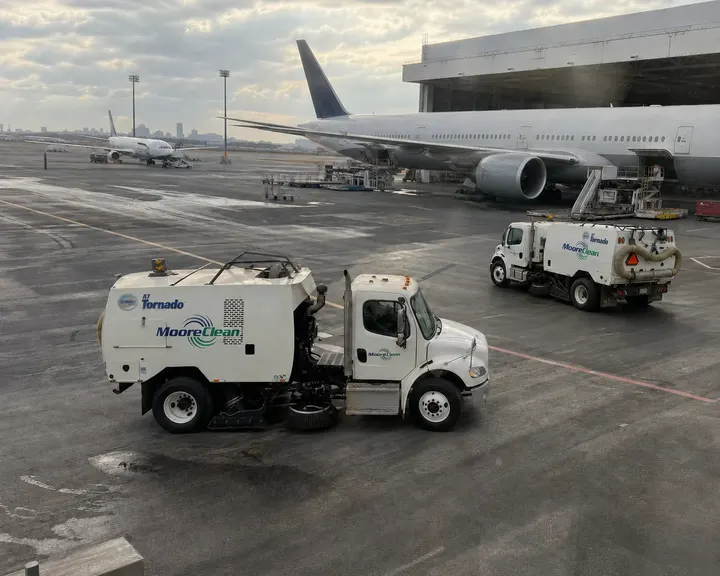 Two Moore Clean A7 Tornado sweepers on an airport apron under a widebody jet's wing at the terminal