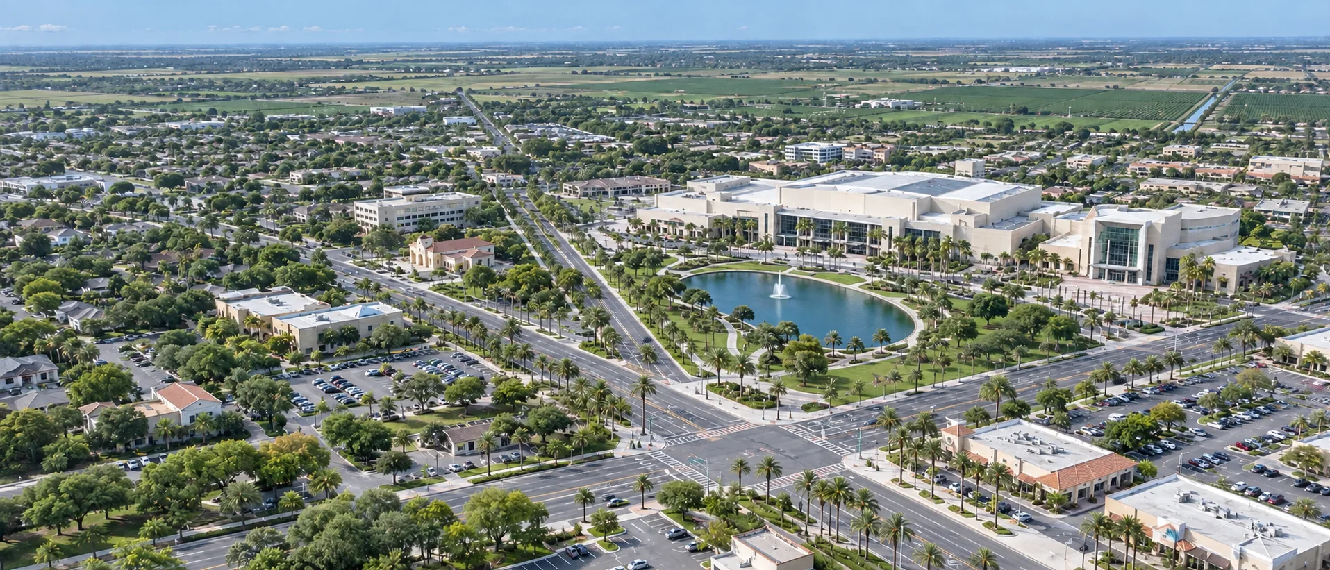 Aerial drone view of McAllen, TX with the convention center, its lake and fountain, palm-lined boulevards, and retail parking lots stretching to Valley farmland