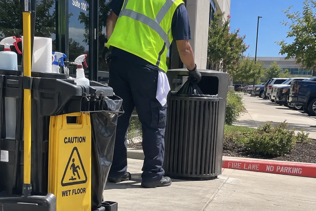 Day porter removing the liner from an exterior trash receptacle beside a janitorial cart